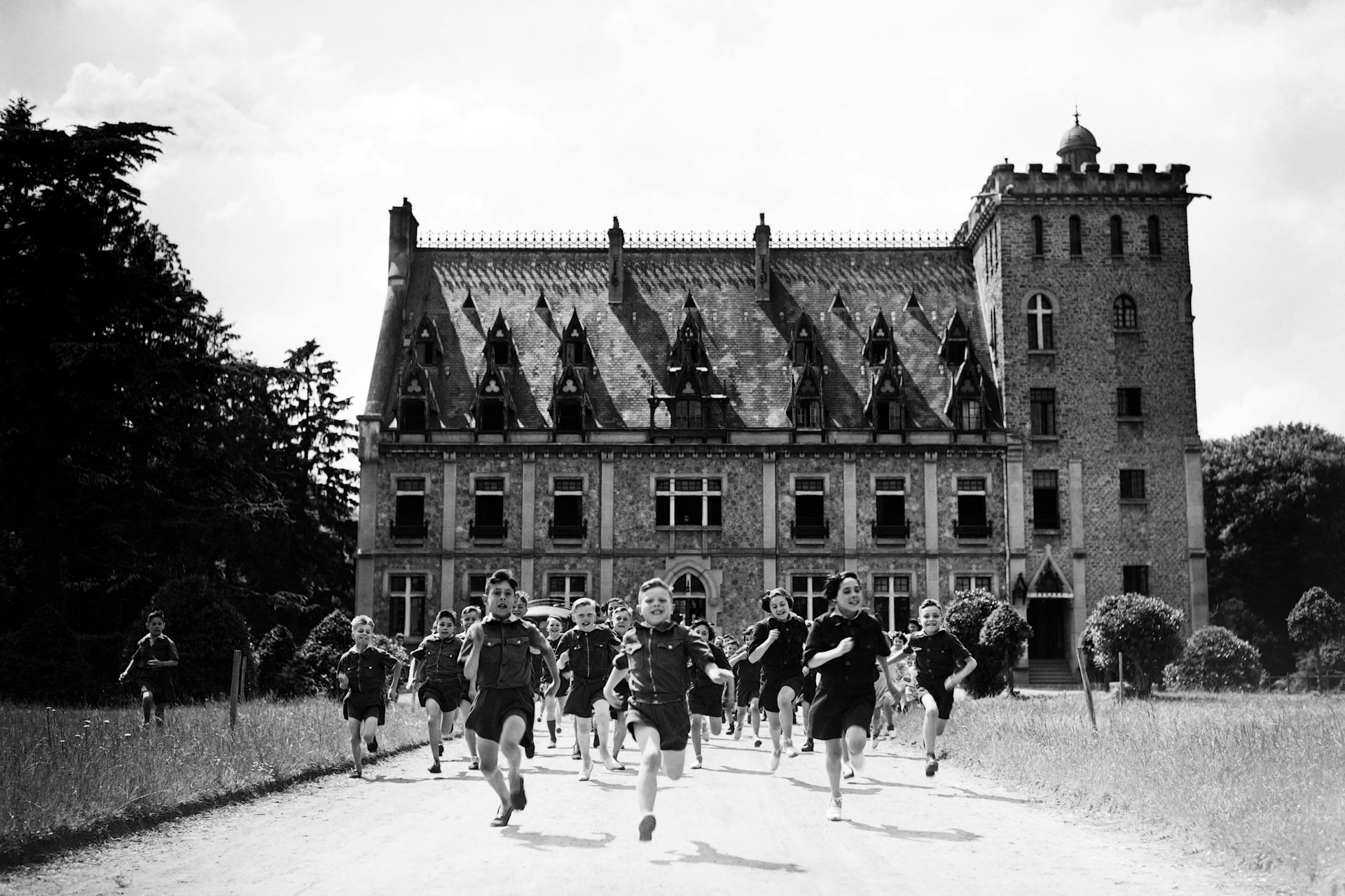 Children wearing uniform shirts and shorts are running towards the viewer in front of a small château.