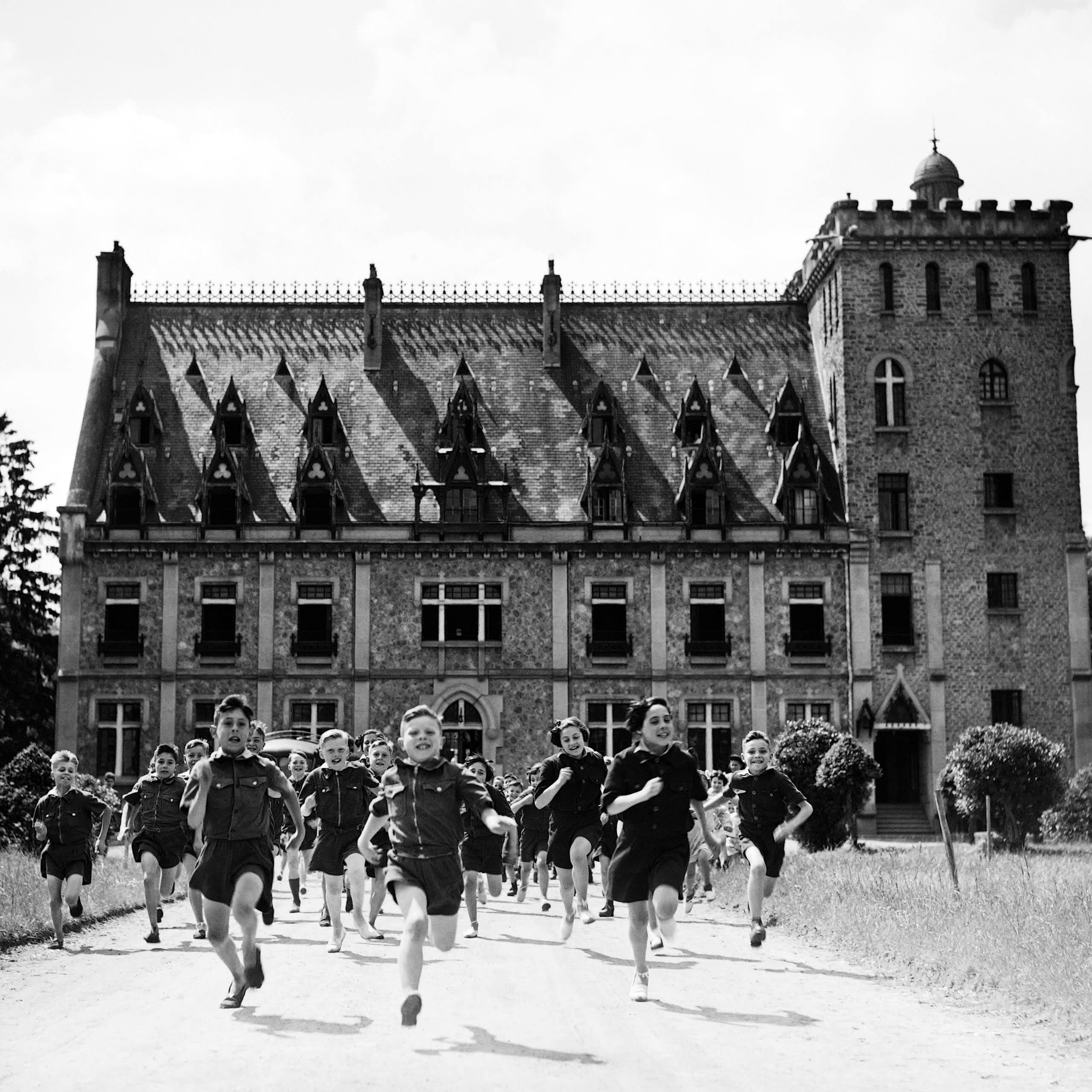 Children wearing uniform shirts and shorts are running towards the viewer in front of a small château.