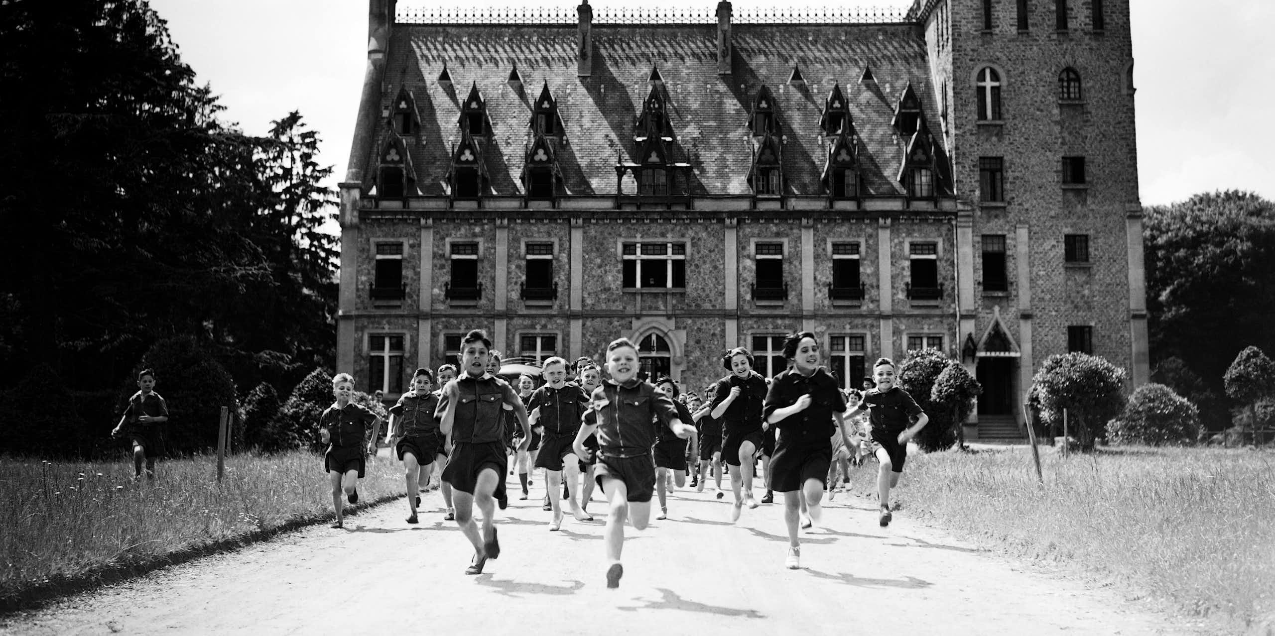 Children wearing uniform shirts and shorts are running towards the viewer in front of a small château.