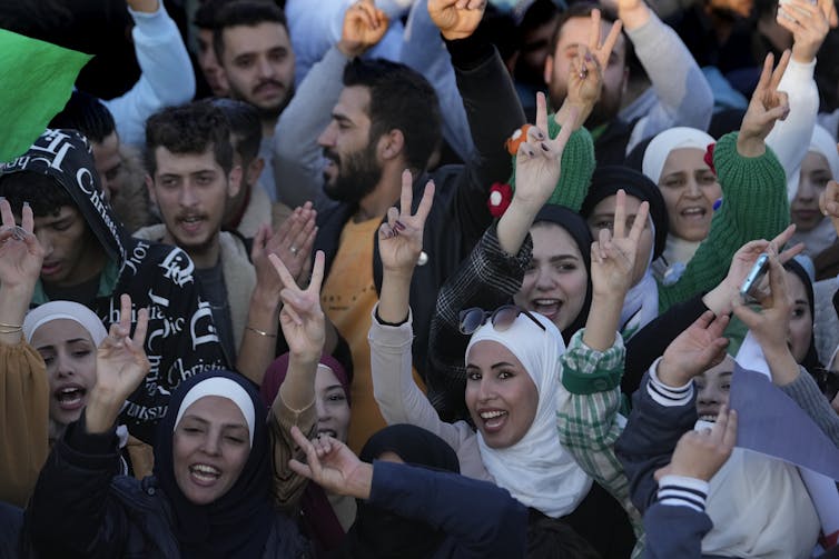 Men and women celebrate in the town square, smiling and holding up V signs.