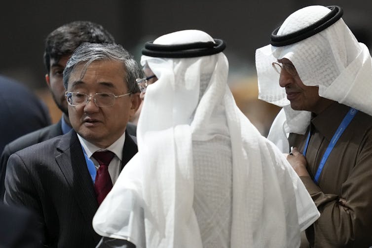 An Asian man stands next to two Saudi officials wearing white scarves.