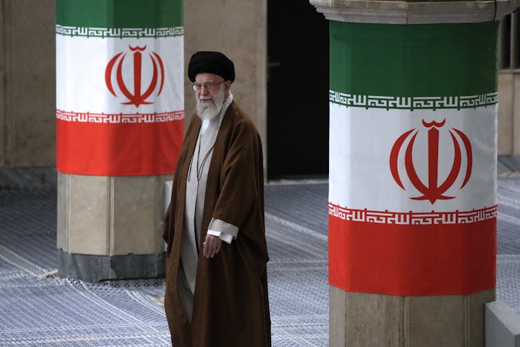 A bearded man in clerical robes walks between two pillars decorated with green, white and red flags.
