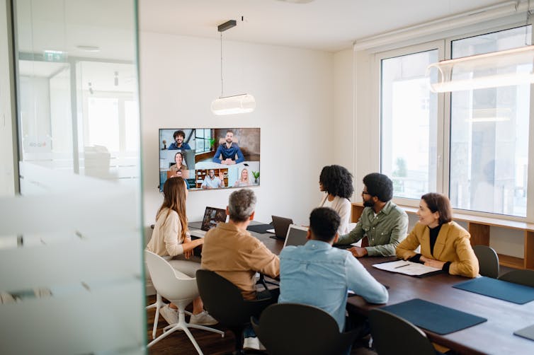 Un groupe de personnes assises à une table de conférence regarde un écran fixé au mur affichant un appel vidéo avec plusieurs personnes