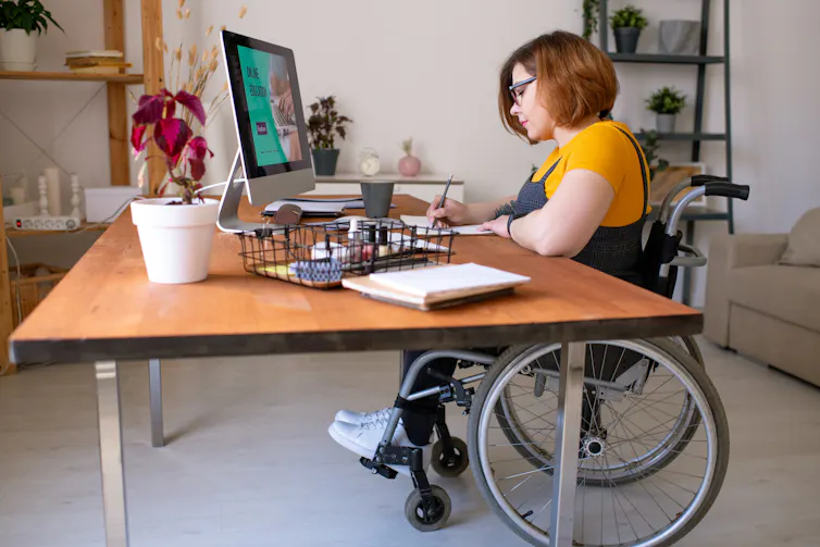 A woman in a wheelchair writes on a pad of paper at a desk