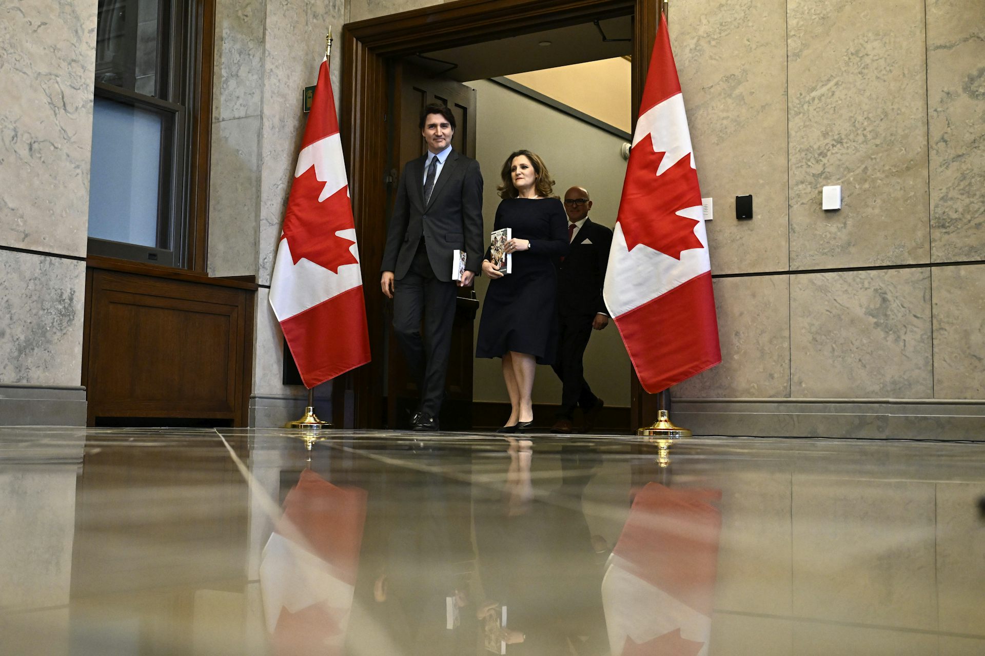 Un homme brun et une femme plus petite aux cheveux blond foncé marchent entre deux drapeaux canadiens