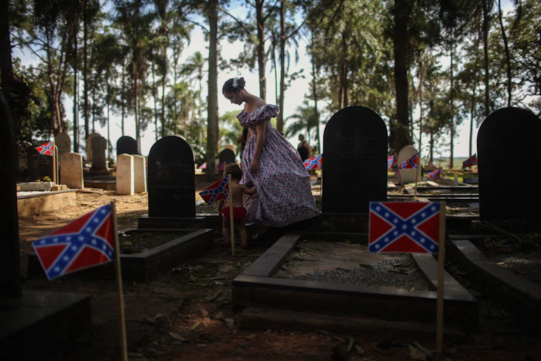 A woman dressed in traditional hoop skirt walks with a child past graves marked with Confederate flags in the American cemetery