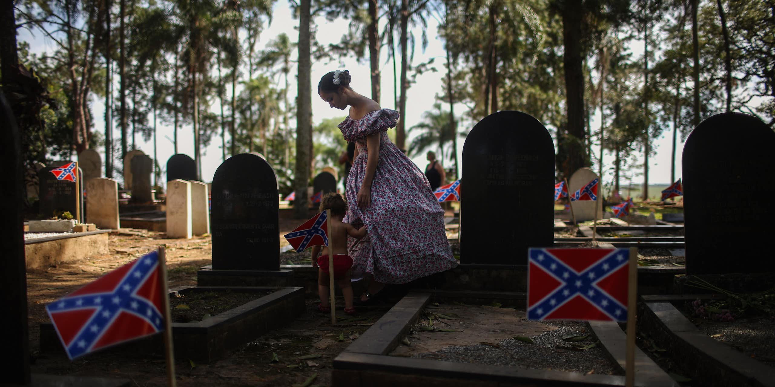A woman dressed in traditional hoop skirt walks with a child past graves marked with Confederate flags in the American cemetery