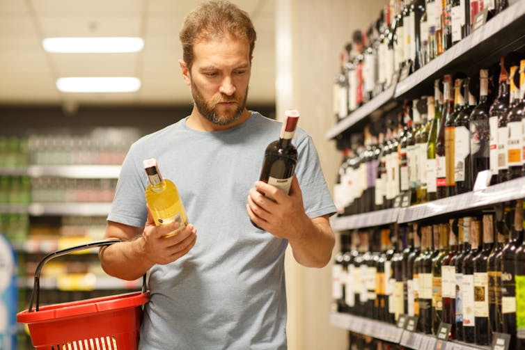 A man holding a shopping basket comparing two bottles in the wine aisle