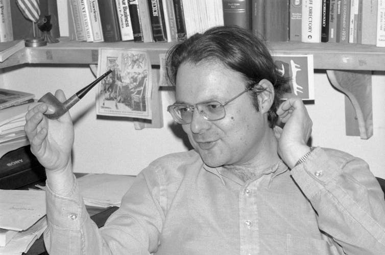 A man with long hair and glasses sitting in front of a bookshelf with a pipe in his hand.