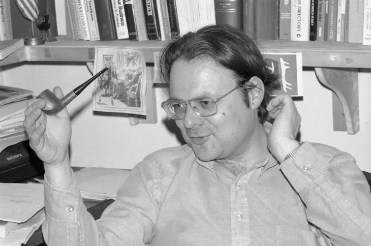 A man with long hair and glasses sitting in front of a bookshelf with a pipe in his hand.