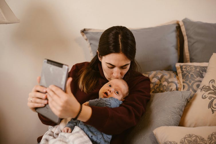 A woman kisses a baby on the head, while holding a tablet in front of her.