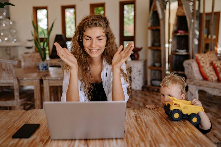 A woman looks at a laptop clapping, a child hovers close to her with a truck