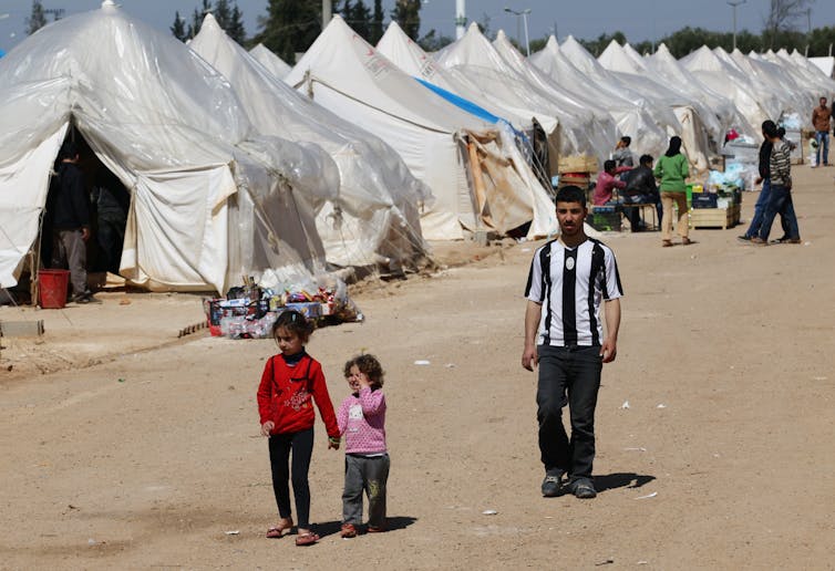 A man and two children walk next to a row of tent encampments.