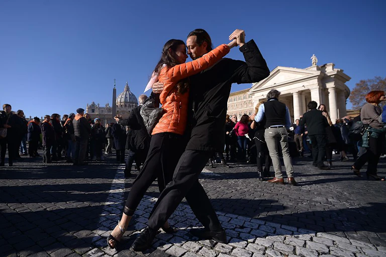 A woman in an orange jacket and a man in a black suit dance in a cobblestone plaza.