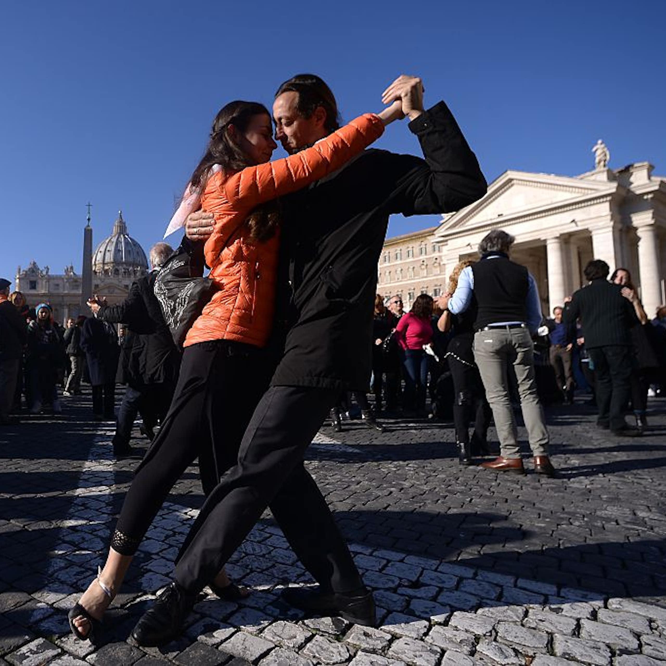 A woman in an orange jacket and a man in a black suit dance in a cobblestone plaza.