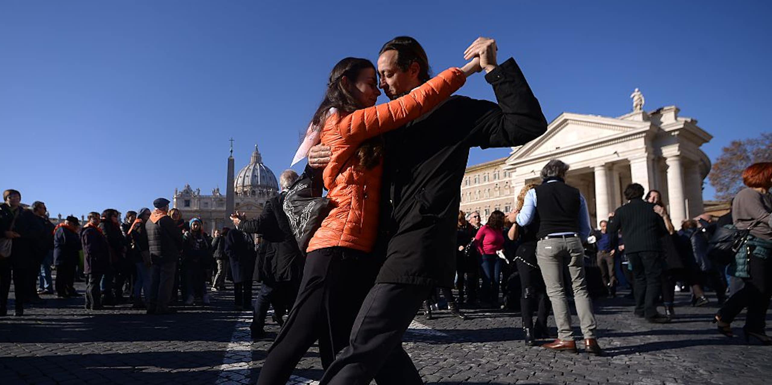 A woman in an orange jacket and a man in a black suit dance in a cobblestone plaza.