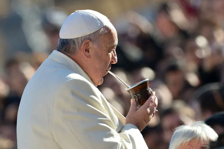 A man in a white jacket and white skullcap sips from a straw coming out of a brown container, with a crowd in the background.