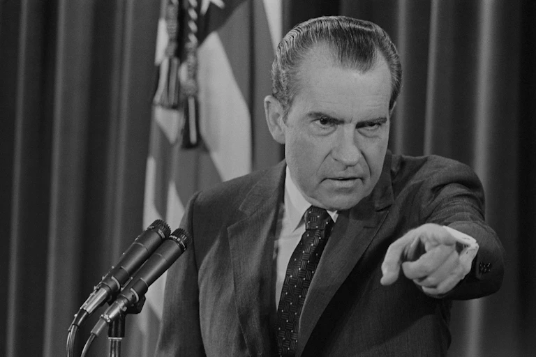 A man with dark receding hair, heavy eyebrows and pointing his finger at someone.