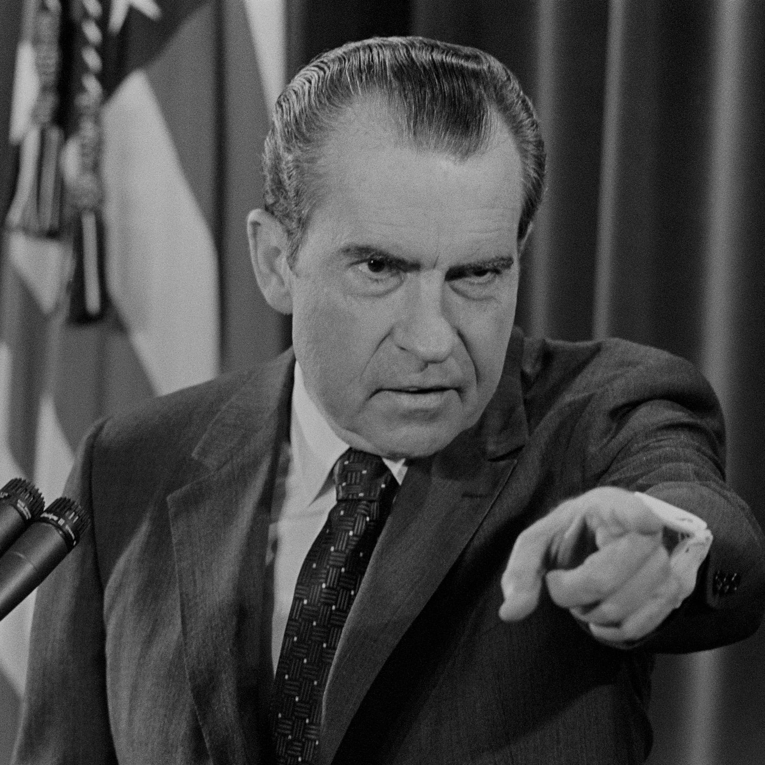 A man with dark receding hair, heavy eyebrows and pointing his finger at someone.