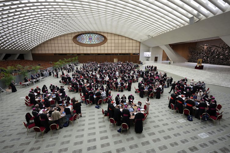 A photograph of a spacious, light-filled room with people, many of them men in black outfits, clustered around round tables.