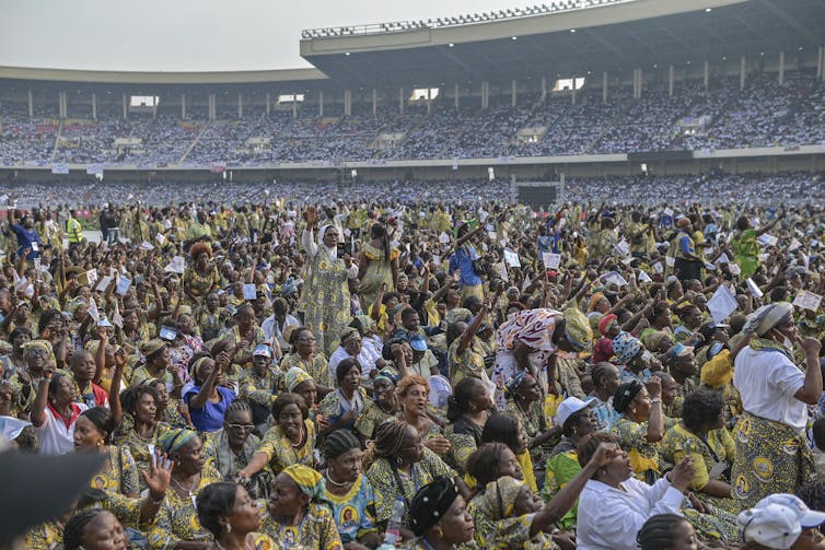 A sports arena filled with people, many of them sitting on the field in yellow- and green-patterned clothing.