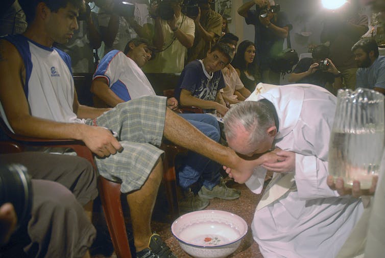 An older man in a white robe kneels next to a bowl of water and kisses the foot of a seated young man in a tank top and striped shorts.