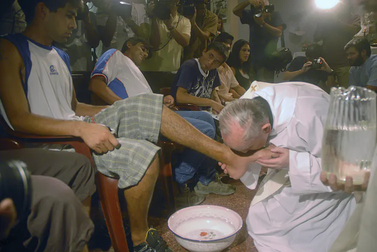 An older man in a white robe kneels next to a bowl of water and kisses the foot of a seated young man in a tank top and striped shorts.