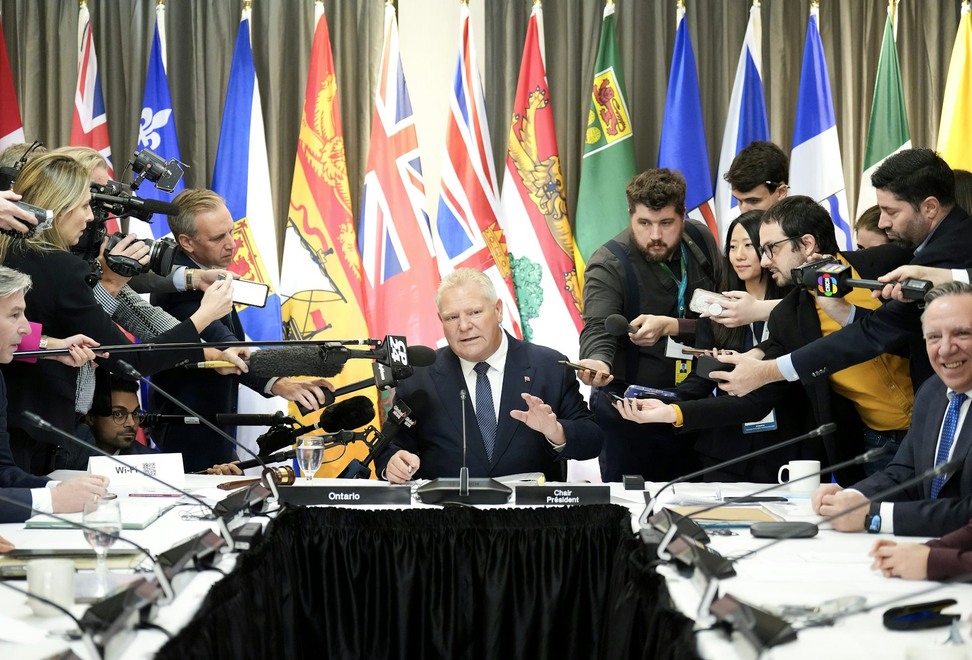 A rotund man with thinning blond-grey hair sits at the head of a table as reporters surround him. A row provincial flags are behind him.