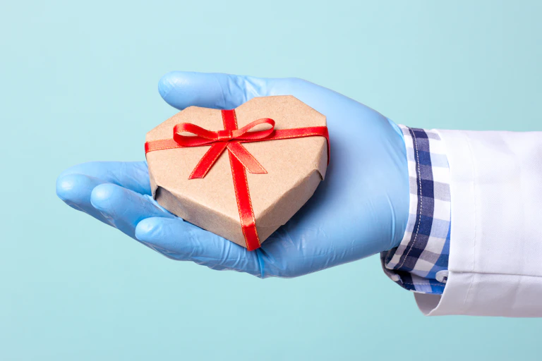hand with blue glove holding wrapped heart-shaped present with red ribbon
