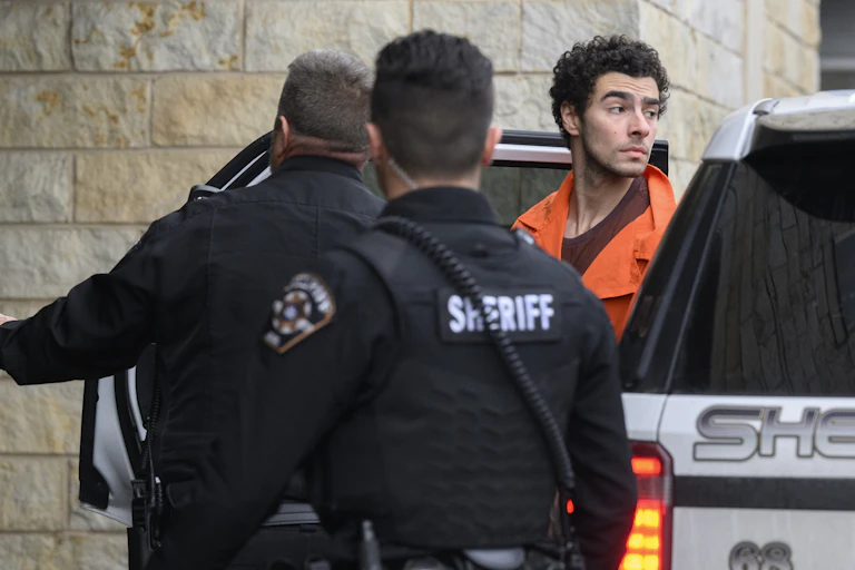 A man wearing an orange top is seen exiting a car. Two men in black stand in front of him.
