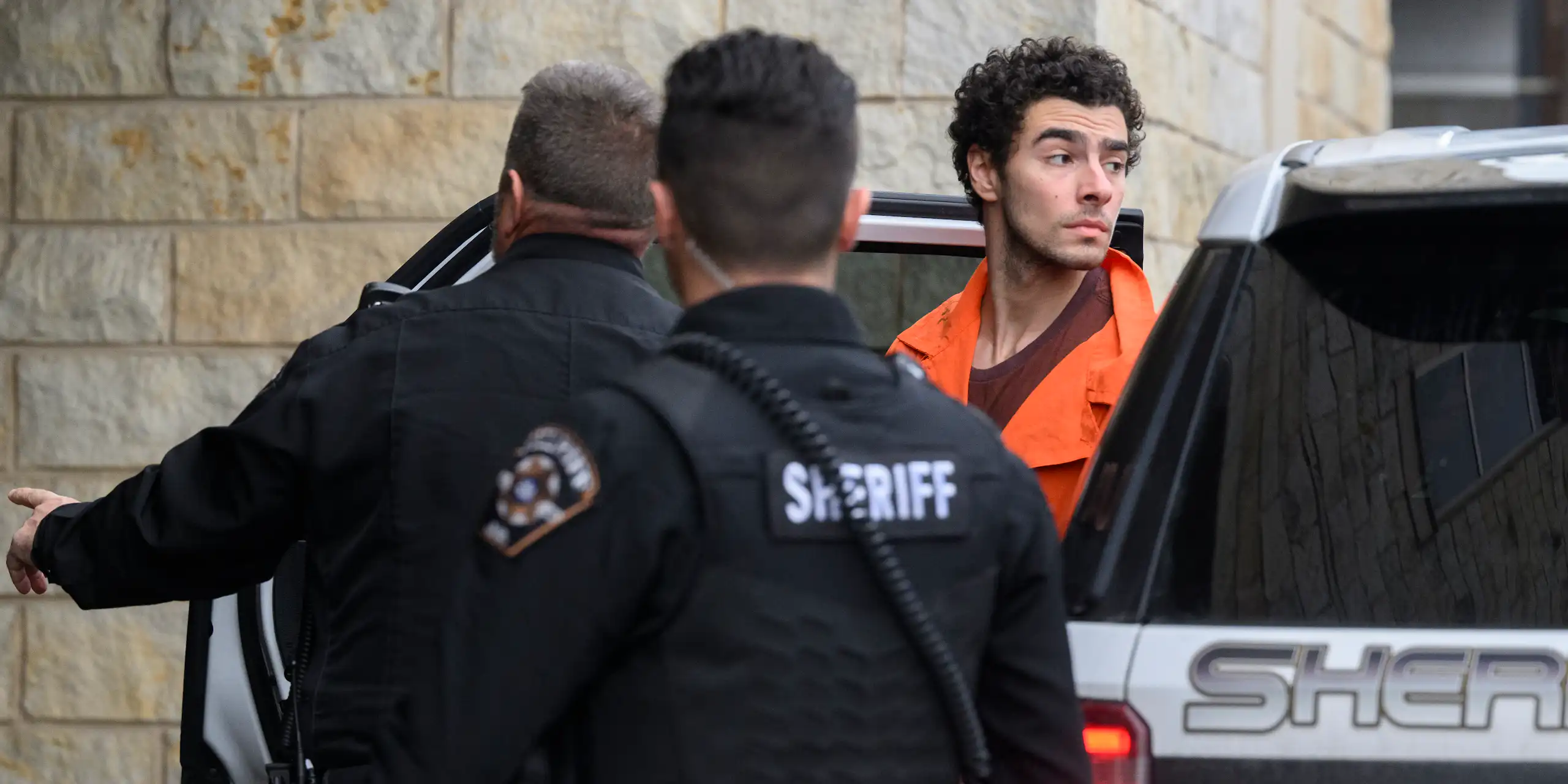 A man wearing an orange top is seen exiting a car. Two men in black stand in front of him.