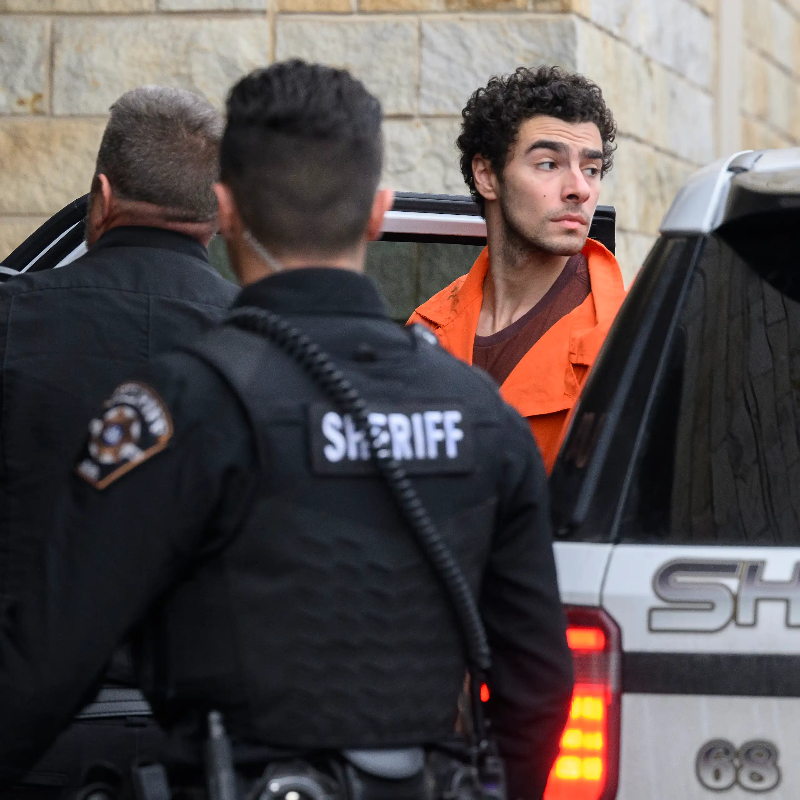 A man wearing an orange top is seen exiting a car. Two men in black stand in front of him.