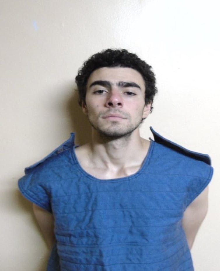 A white man with dark hair wears a blue shirt, with his hands behind his back, and looks at the camera.