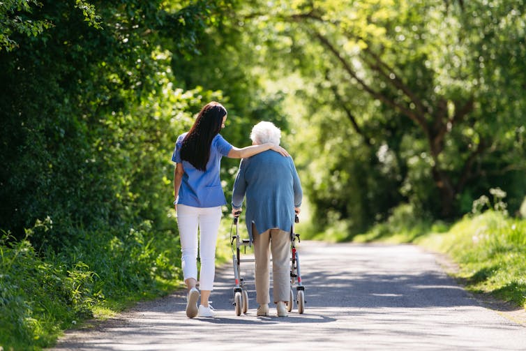 A young woman walks with her arm around the shoulder of an elderly woman who is using a walker