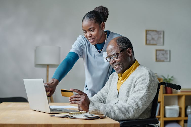 An elderly man seated at a table looks at a banking card while a young woman stands over his shoulder and points at the screen of an open laptop