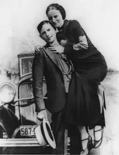A black-and-white photo shows a young woman and man smiling as they stand against a car.