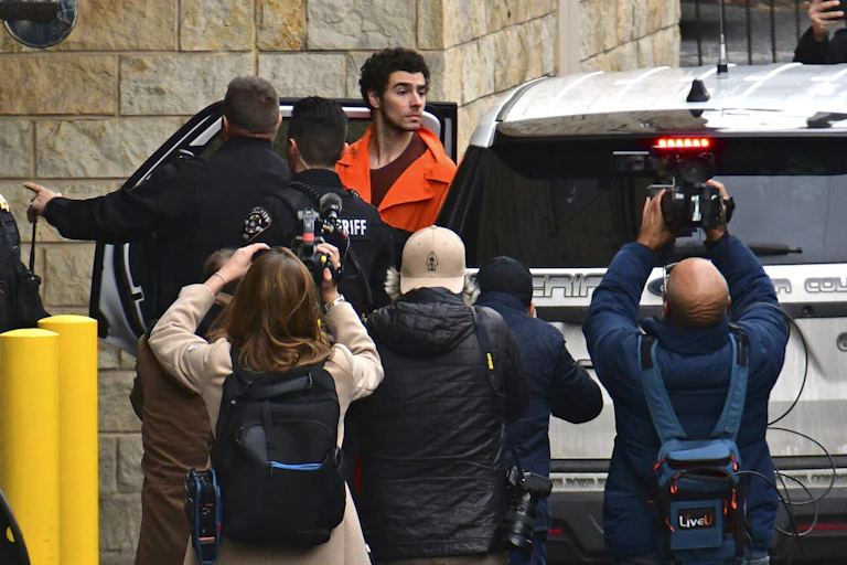 A young man with dark hair in an orange prison jumpsuit exits a police vehicle amid a media throng.