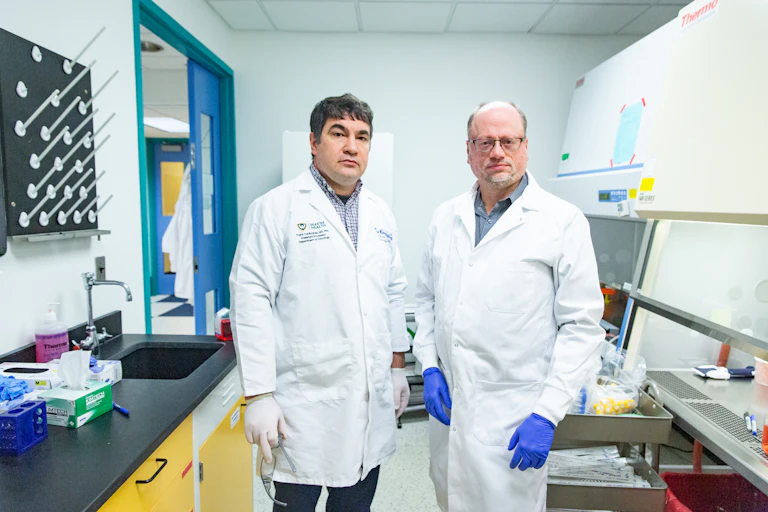 Two men wearing lab coats and gloves standing in a lab with stern looks on their faces