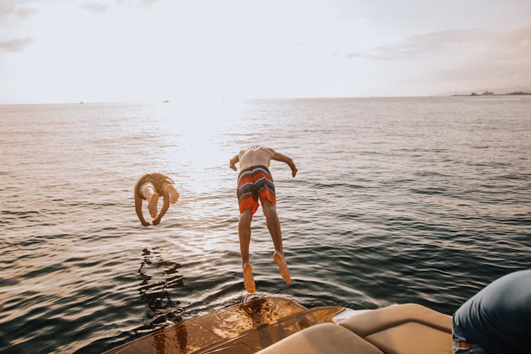 Two young men jump from a boat into calm water, the sun setting on the horizon.