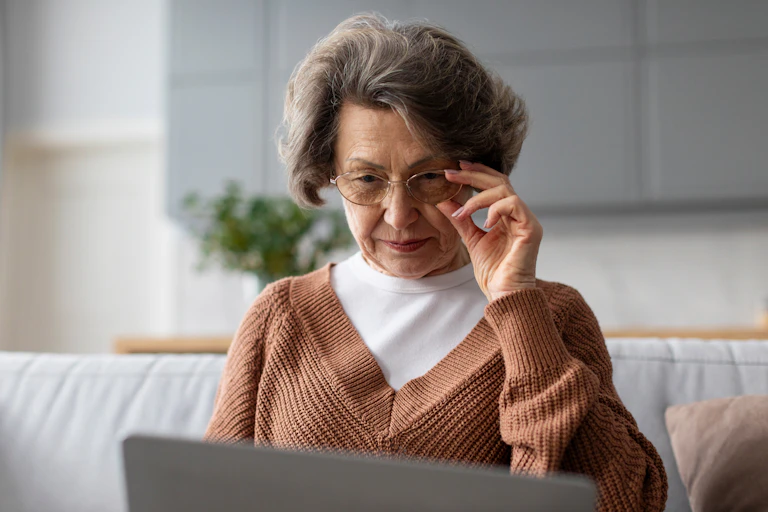 An elderly woman adjusts her glasses while looking at the screen of an open laptop