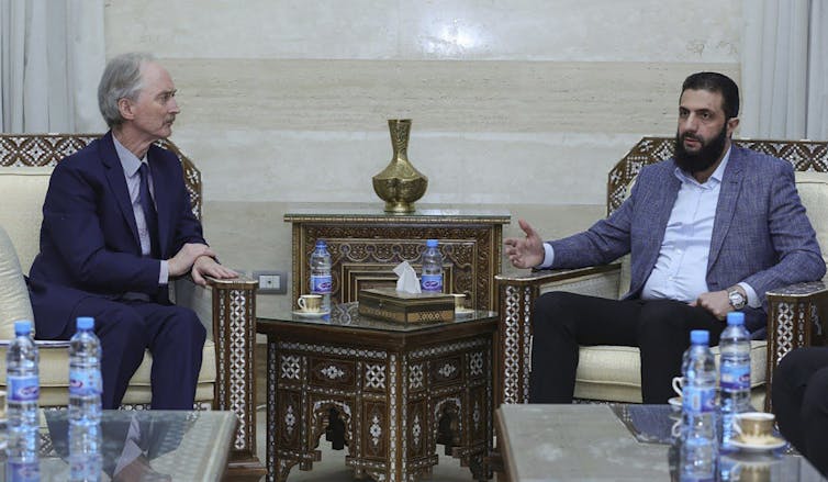 A white man in a suit sits across from a bearded Middle Eastern man in a blazer and dress shirt