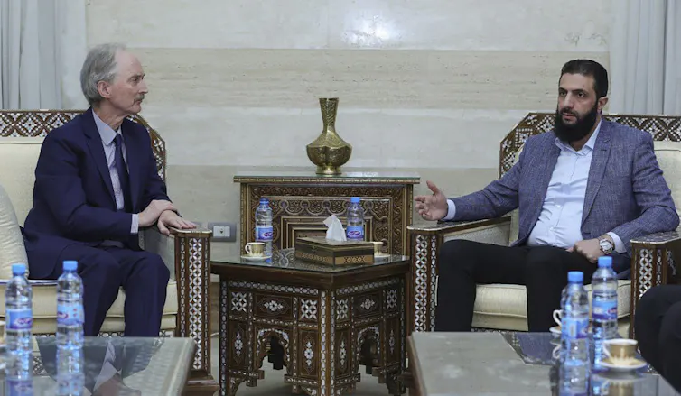 A white man in a suit sits across from a bearded Middle Eastern man in a blazer and dress shirt