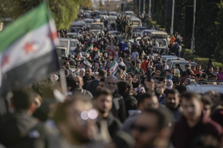A massive crowd of people walk down a car-lined street carrying Syrian flags