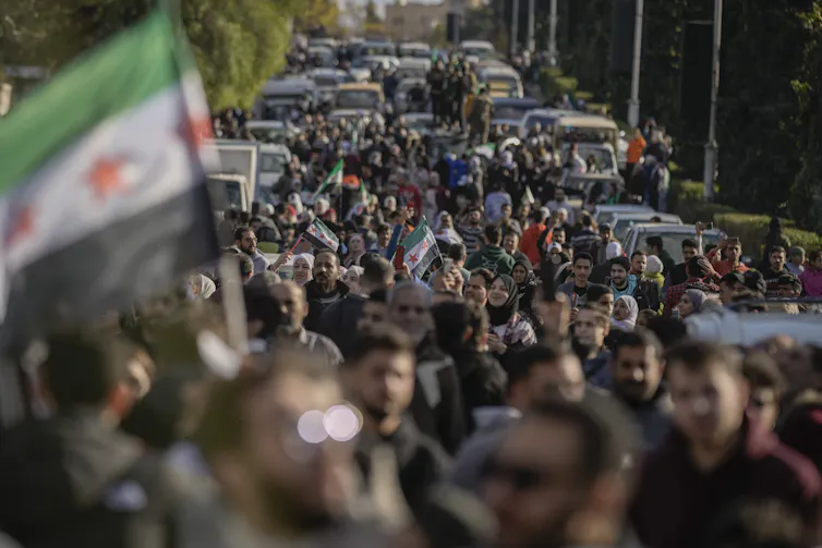 A massive crowd of people walk down a car-lined street carrying Syrian flags
