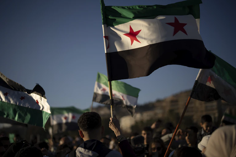Syrian flags fly against a blue sky, held in the hands of people marching together