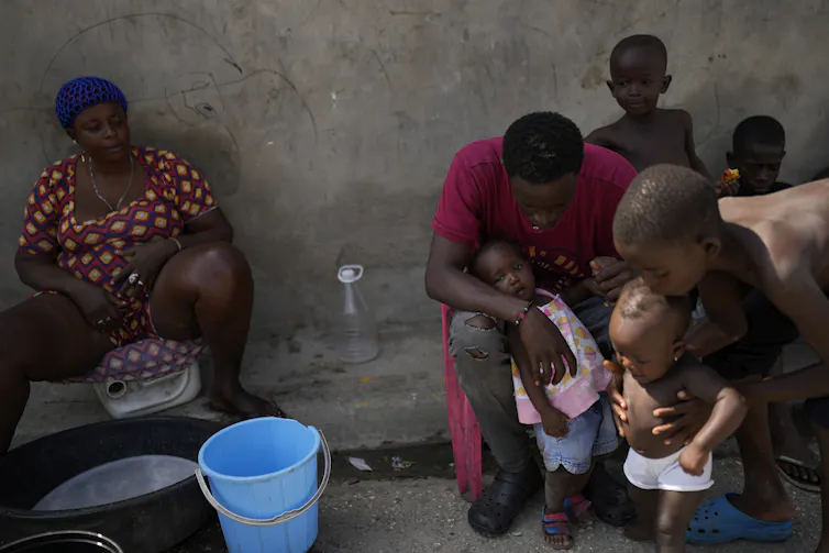 A Black family with several small children sit in the shade outside a concrete structure.