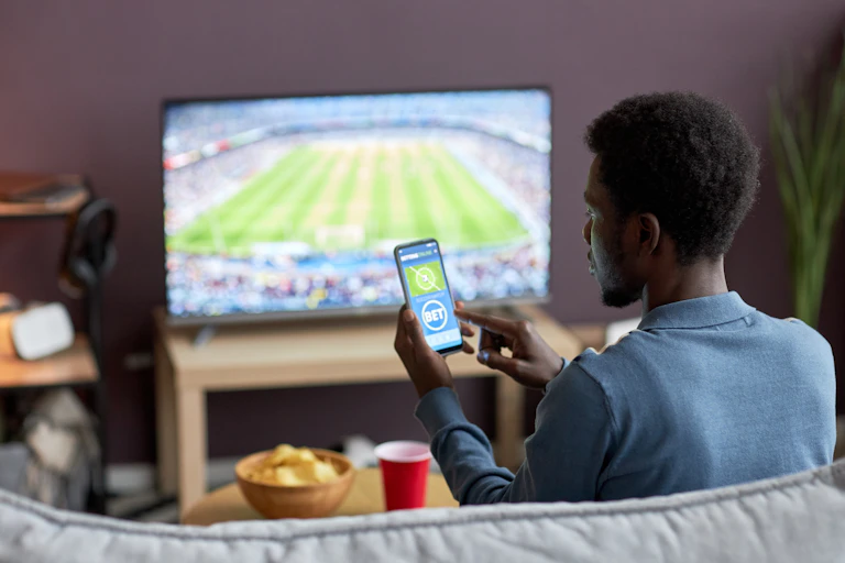 A man watching a sports event on television and placing a bet on his phone