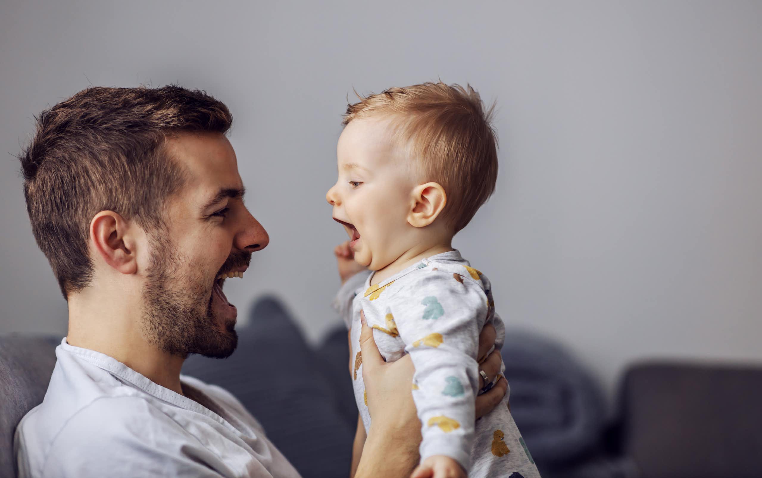 A man holding up a baby, both have their mouths open talking to each other