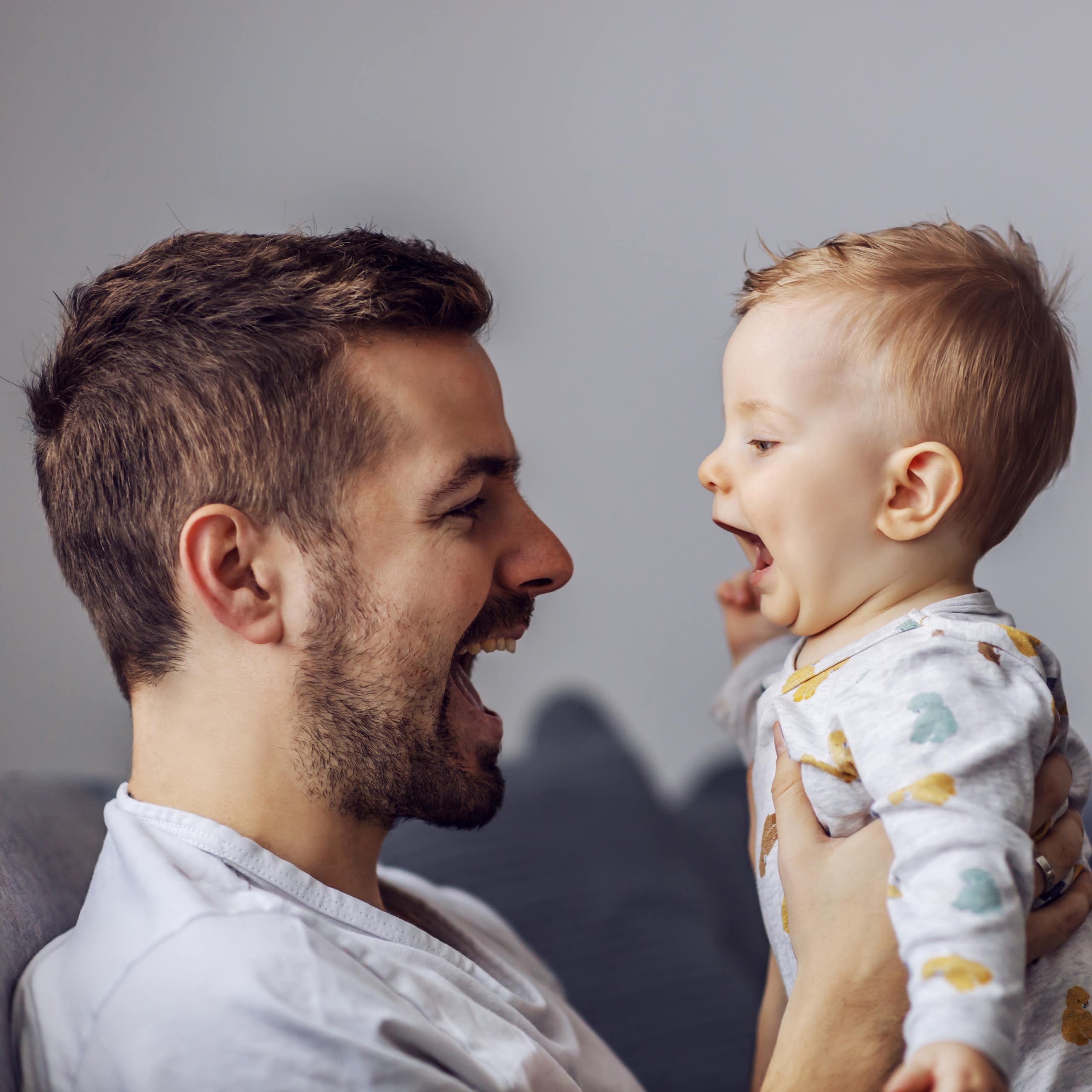 A man holding up a baby, both have their mouths open talking to each other