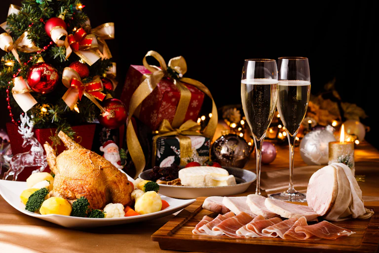 Un table de repas bien remplie avec de la charcuterie, une volaille, du fromage, des coupes de champagne, un petit sapin artificiel et des cadeaux.